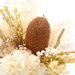 A mix of preserved flowers and silk flowers of hydrangea, misty, viburnum, bunny tails, rice flower, baby’s breath, peony flower, banksia and palm in a in white vase.