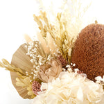 A mix of preserved flowers and silk flowers of hydrangea, misty, viburnum, bunny tails, rice flower, baby’s breath, peony flower, banksia and palm in a in white vase.