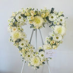Heart-shaped floral arrangement with white and green flowers on a stand against a light gray background.