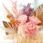 A mix of preserved flowers and silk flowers of hydrangea, Italian ruscus, billy button, misty, peony, bunny tail and palm in a in pink vase.