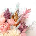 A mix of preserved flowers and silk flowers of hydrangea, Italian ruscus, billy button, misty, peony, bunny tail and palm in a in pink vase.