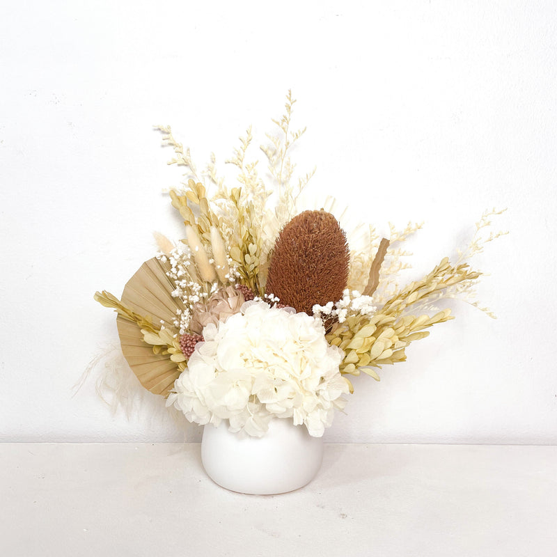 A mix of preserved flowers and silk flowers of hydrangea, misty, viburnum, bunny tails, rice flower, baby’s breath, peony flower, banksia and palm in a in white vase.