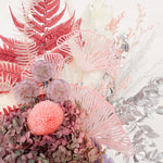 A mix of preserved flowers and silk flowers of hydrangea, ruscus, chrysanthemum, amaranthus, cloudy leaf, tiki grass, leather fern and scabiosa in a in white vase.