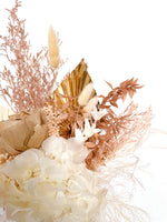 A mix of preserved flowers and silk flowers of hydrangea, misty, nigella, bunny tails, rice flower, baby’s breath, peony flower and palm in a in white vase.