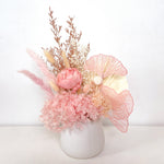 A mix of preserved flowers and silk flowers of hydrangea, Italian ruscus, bunny tails, baby's breath, rice flower, peony flower, palm and fluffy stick in a in white vase.