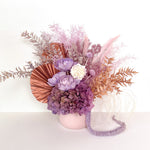 A mix of preserved flowers and silk flowers of hydrangea, Italian ruscus, billy button, fox tail, peony, tiki fern and palm arranged in a in pink vase.