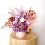 A mix of preserved flowers and silk flowers of hydrangea, Italian ruscus, billy button, fox tail, peony, tiki fern and palm arranged in a in pink vase.