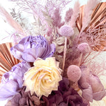 A mix of preserved flowers and silk flowers of hydrangea, Italian ruscus, billy button, fox tail, peony, tiki fern and palm arranged in a in pink vase.