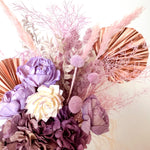 A mix of preserved flowers and silk flowers of hydrangea, Italian ruscus, billy button, fox tail, peony, tiki fern and palm arranged in a in pink vase.