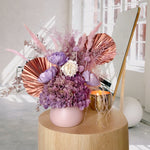 A mix of preserved flowers and silk flowers of hydrangea, Italian ruscus, billy button, fox tail, peony, tiki fern and palm arranged in a in pink vase.