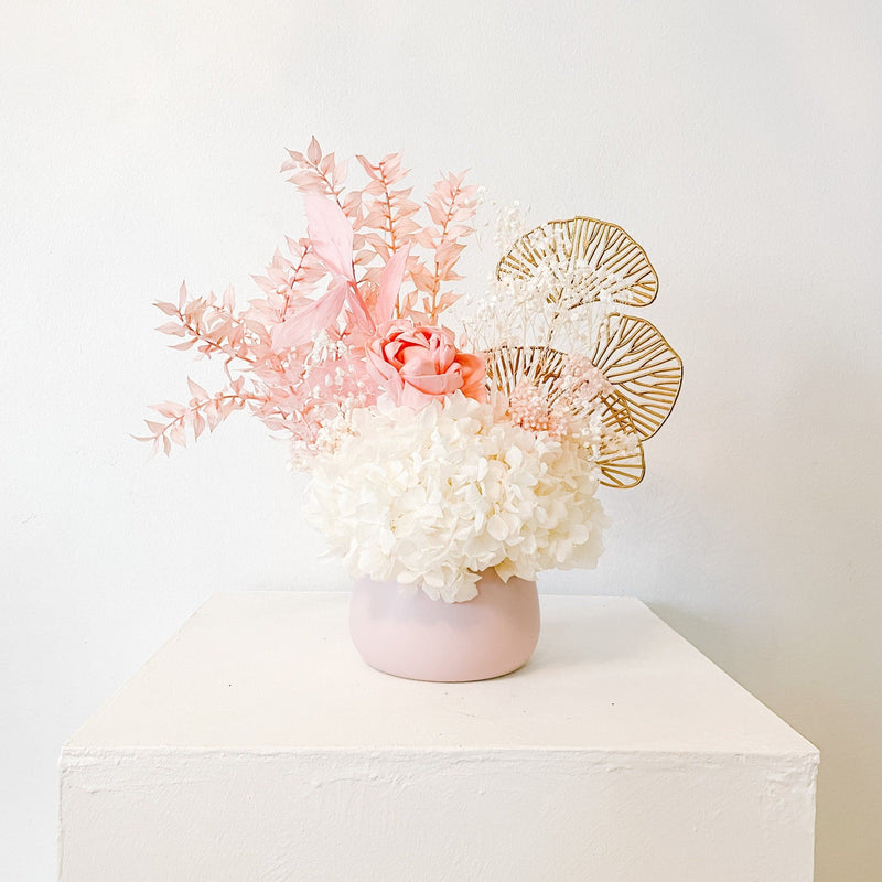 A mix of preserved flowers and silk flowers of hydrangea, Italian ruscus, gingko, baby's breath, rice flower, and peony flower arranged in a in pink vase. 