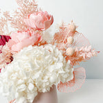 A mix of preserved flowers and silk flowers of hydrangea, Italian ruscus, billy button, amaranthus, misty, peony and palm arranged in a in pink vase.