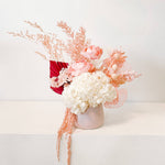 A mix of preserved flowers and silk flowers of hydrangea, Italian ruscus, billy button, amaranthus, misty, peony and palm arranged in a in pink vase.