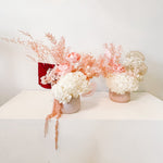A mix of preserved flowers and silk flowers of hydrangea, Italian ruscus, gingko, baby's breath, rice flower, and peony flower arranged in a in pink vase.