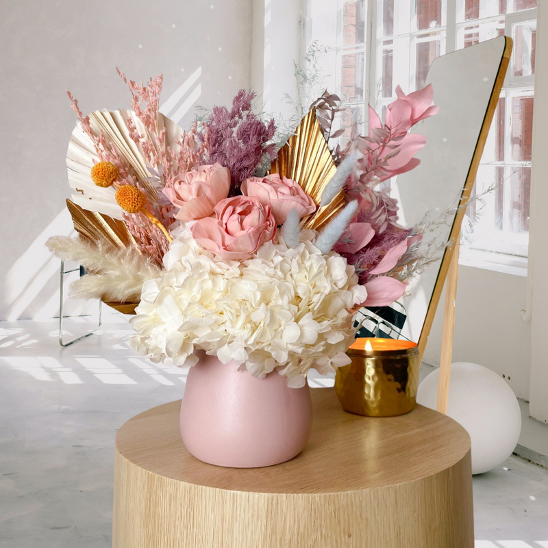 A mix of preserved flowers and silk flowers of hydrangea, Italian ruscus, billy button, misty, peony, bunny tail and palm in a in pink vase.
