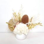 A mix of preserved flowers and silk flowers of hydrangea, misty, viburnum, bunny tails, rice flower, baby’s breath, peony flower, banksia and palm in a in white vase.