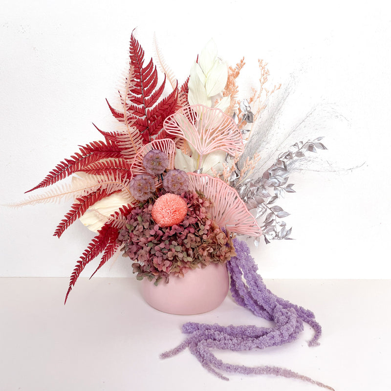 A mix of preserved flowers and silk flowers of hydrangea, ruscus, chrysanthemum, amaranthus, cloudy leaf, tiki grass, leather fern and scabiosa in a in white vase.