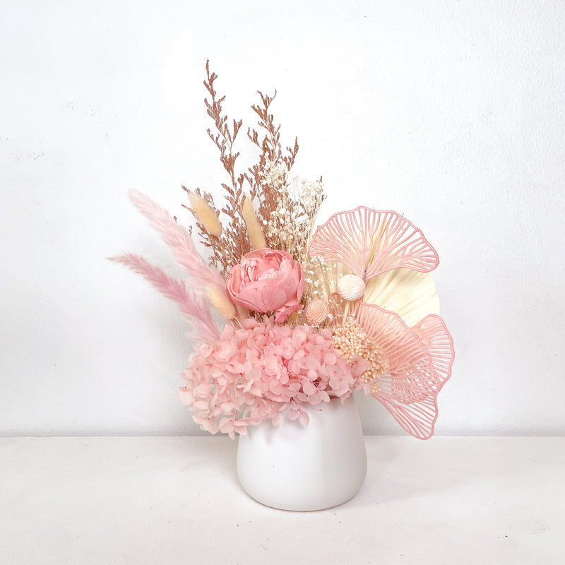 A mix of preserved flowers and silk flowers of hydrangea, Italian ruscus, bunny tails, baby's breath, rice flower, peony flower, palm and fluffy stick in a in white vase.