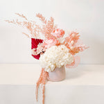 A mix of preserved flowers and silk flowers of hydrangea, Italian ruscus, billy button, amaranthus, misty, peony and palm arranged in a in pink vase. 