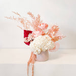 A mix of preserved flowers and silk flowers of hydrangea, Italian ruscus, billy button, amaranthus, misty, peony and palm arranged in a in pink vase. 