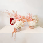 A mix of preserved flowers and silk flowers of hydrangea, Italian ruscus, billy button, amaranthus, misty, peony and palm arranged in a in pink vase. 