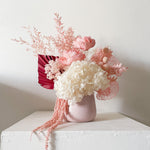 A mix of preserved flowers and silk flowers of hydrangea, Italian ruscus, billy button, amaranthus, misty, peony and palm arranged in a in pink vase. 