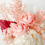 A mix of preserved flowers and silk flowers of hydrangea, Italian ruscus, billy button, amaranthus, misty, peony and palm arranged in a in pink vase. 