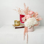 A mix of preserved flowers and silk flowers of hydrangea, Italian ruscus, billy button, amaranthus, misty, peony and palm arranged in a in pink vase. 