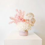 A mix of preserved flowers and silk flowers of hydrangea, Italian ruscus, gingko, baby's breath, rice flower, and peony flower arranged in a in pink vase. 