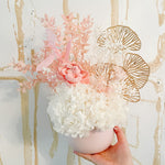 A mix of preserved flowers and silk flowers of hydrangea, Italian ruscus, gingko, baby's breath, rice flower, and peony flower arranged in a in pink vase. 